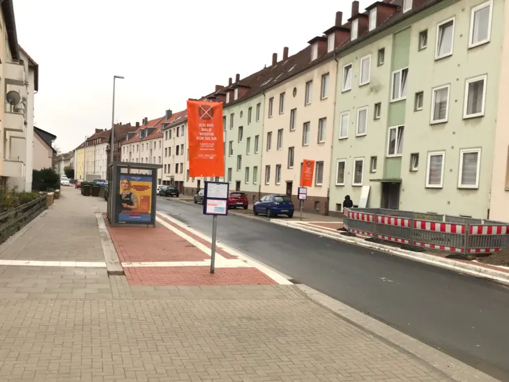 Zwischen zwei Hausfronten verläuft eine asphaltierte Straße. Links auf dem Gehweg steht ein verdecktes Haltestellenschild, dahinter ein Warteunterstand. Auf der Fläche verlaufen weiße Linien.