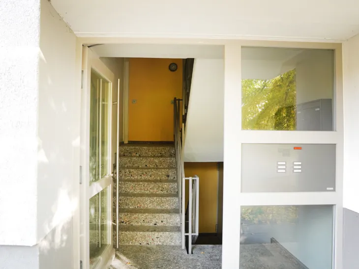 Eingang in ein Mehrparteienhaus durch eine Tür mit hellen Streben und Balken und Glasscheiben.
