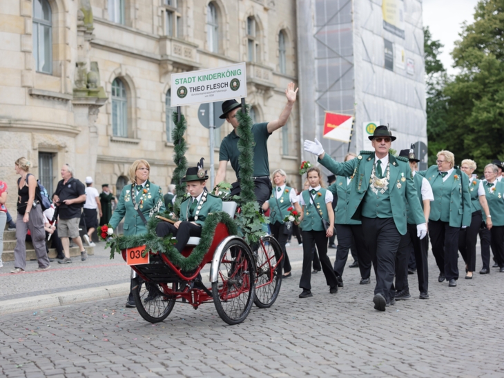 Schützen und Schützinnen auf einer Straße. Der Kinderkönig wird in einem Wagen gefahren.