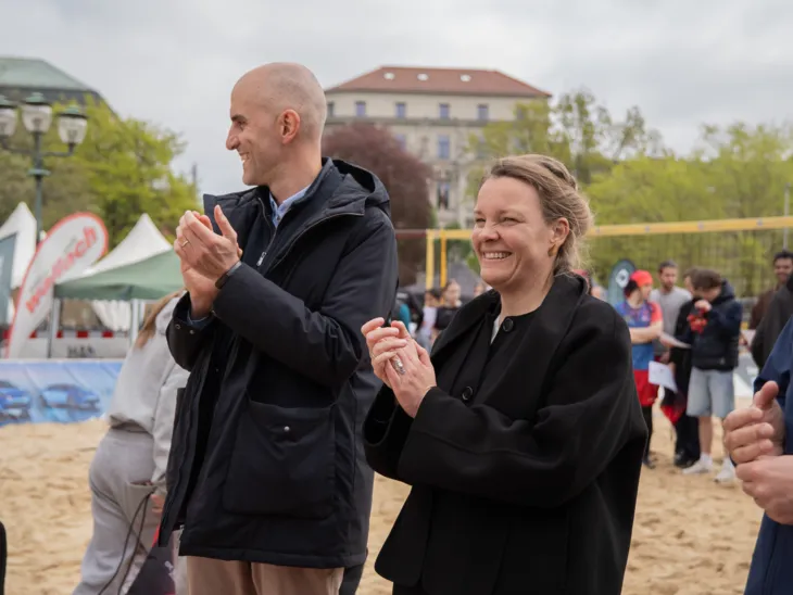 Links ein Mann und rechts eine Frau, die klatschen. Im Hintergrund ein Beachvolleyballfeld.