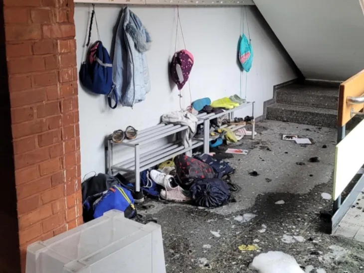 An einer Wandleiste hängen drei Turnbeutel und eine Sweatjacke, darunter auf und neben einer Bank liegen Schuhe, Taschen und Kleidung, davor eine große Pfütze mit weißem Schaum und einer umgestülpten transparenten Kiste.