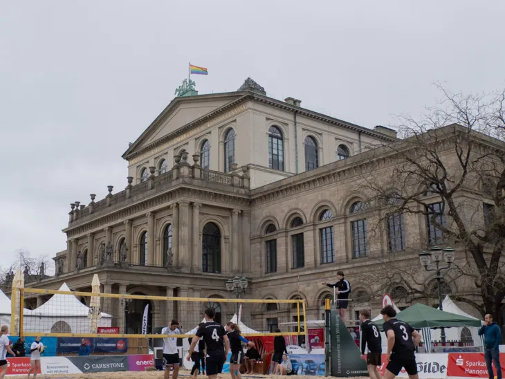 Ein Beachvolleyball-Feld mit Spieler*innen darauf, im Hintergrund ein altes Gebäude, in dem die Hannover Oper ist.