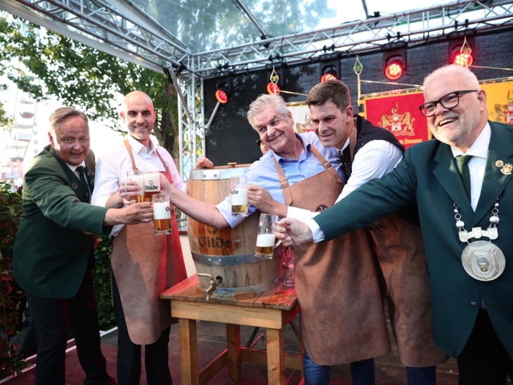 Fünf Männer an einem Bierfass. Sie schwenken die Gläser.