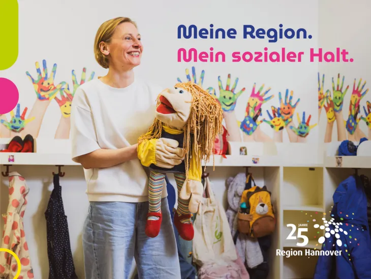 Eine Frau steht in einer KiTa vor der Garderobe mit Kinderjacken und spielt mit einer Handpuppe.