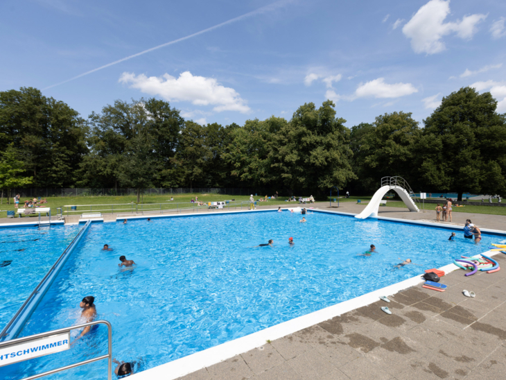 Freibadbecken mit Trennwand, man sieht den Nichtschwimmerbereich, eine Rutsche und Personen im Wasser
