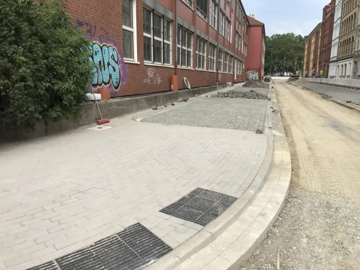Zwischen zwei Gebäuden verläufen eine Straße und ein Fußweg. Die Straße besteht noch aus Sand und Schotter, der Fußweg links endet an der Ecke einer Kreuzung mit eingefassten schwarzen Steinen, auf denen Rillen angeordnet sind.