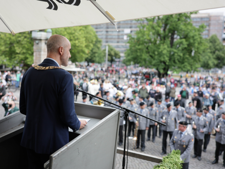 Ein Mann am Redepult. Auf einem Platz zahlreiche Schütz*innenvereine.