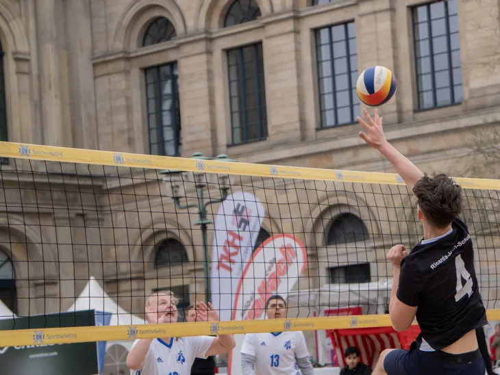 Ein Beachvolleyball fliegt über ein Netz, auf beiden Seiten stehen Spieler und auf der rechten Seite springt ein Spieler zum Ball.