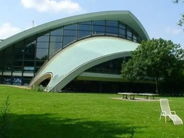 Schwimmhalle, davor grüne Wiese mit zwei weißen Liegestühlen