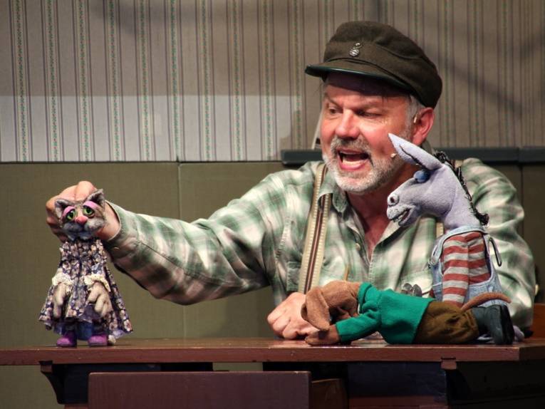 Mann mit einer Katzenpuppe, einer Eselpuppe und einer Hundepuppe.