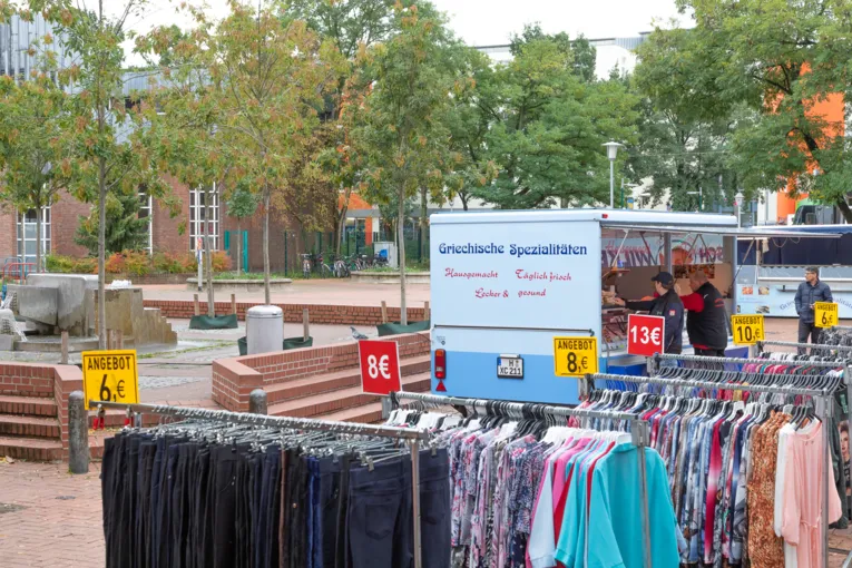 Auf einem Wochenmarktstand werden Kleidungsst&uuml;cke angeboten; im Hintergrund ein Wagen mit griechischer Feinkost.