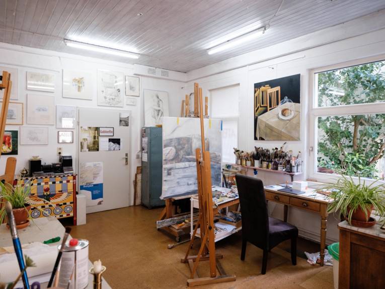 Blick in das Atelier eines Künstlers: Werke hängen an den Wänden, Arbeitsmaterialien wie Pinsel und Staffeleien sind im Raum verteilt.