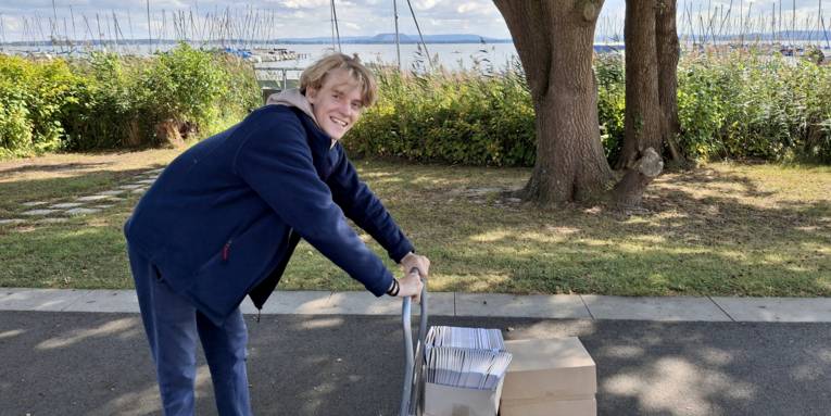 Ein Mann schiebt einen Rollwagen mit Kartons, in den Kartons sind Flyer.