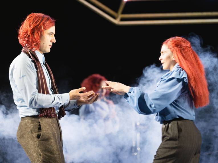 Auf einer Bühne halten sich ein Mann und eine Frau, beide mit roten Haaren, an den Händen. Im Hintergrund ist eine weitere Frau mit roten Haaren und Bühnennebel zu sehen.
