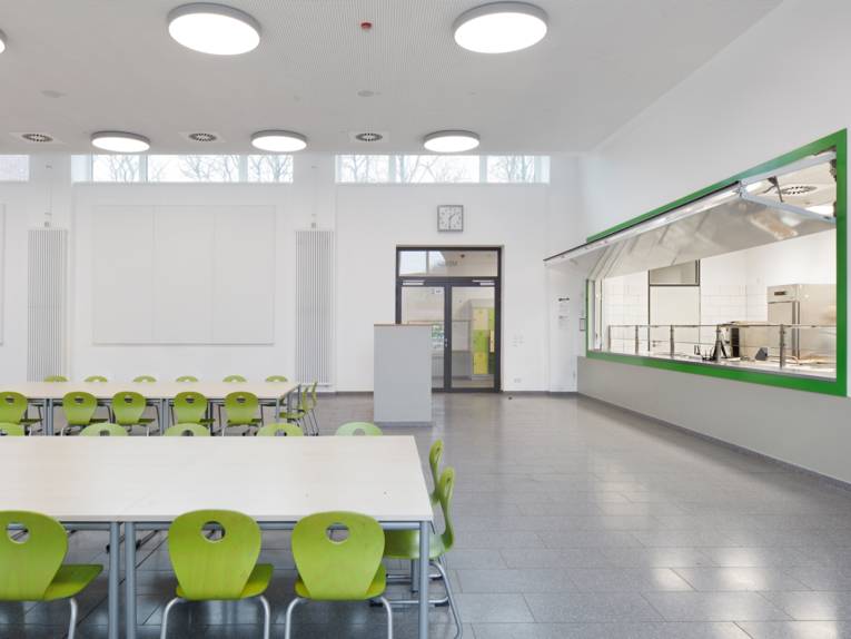 Eine gro&szlig;e Halle, links Tische mit hellgr&uuml;nen St&uuml;hlen, rechts eine Wand mit einem offenen Bereich zur Essensausgabe mit gr&uuml;ner Umrandung.