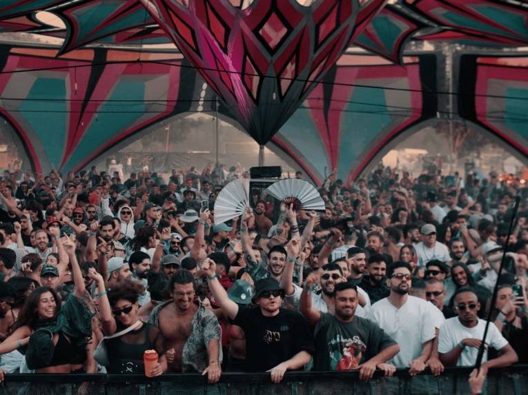 Eine große Menschenmenge feiert vor einer Bühne unter einem farbenfrohen, geometrisch gestalteten Zeltdach. Die dicht gedrängten Festivalbesucherinnen und Festivalbesucher tanzen, heben die Arme in die Luft und schaffen eine lebendige, energiegeladene Atmosphäre.