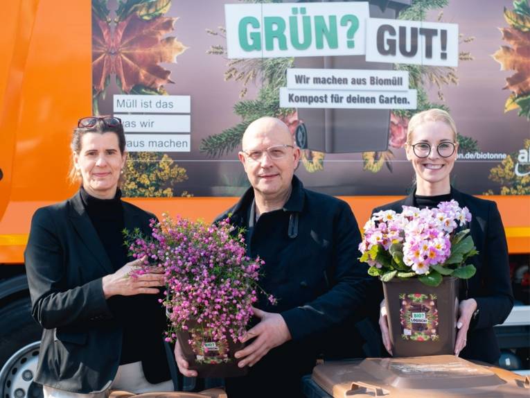 Zwei Frauen und ein Mann stehen vor einem orangenen Müllfahrzeug und halten Blumen mit in die Kamera.