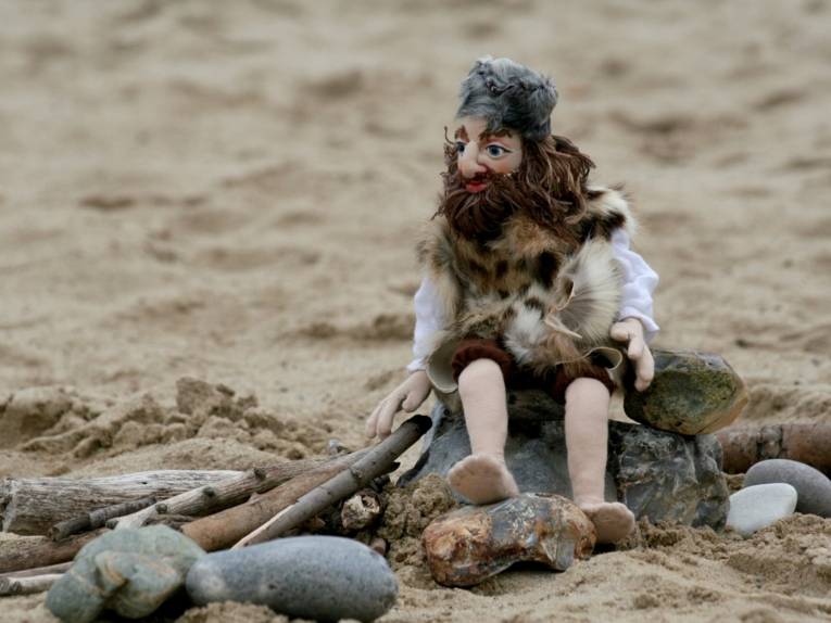 Zu sehen ist eine Marionette in zerlumpter Kleidung und einem langen Bart, die am Strand sitz.