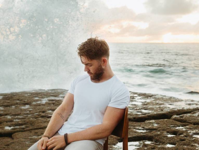 Mann mit Bart und weißem Shirt sitzt auf einem Holzstuhl an der Küste, im Hintergrund das Meer.