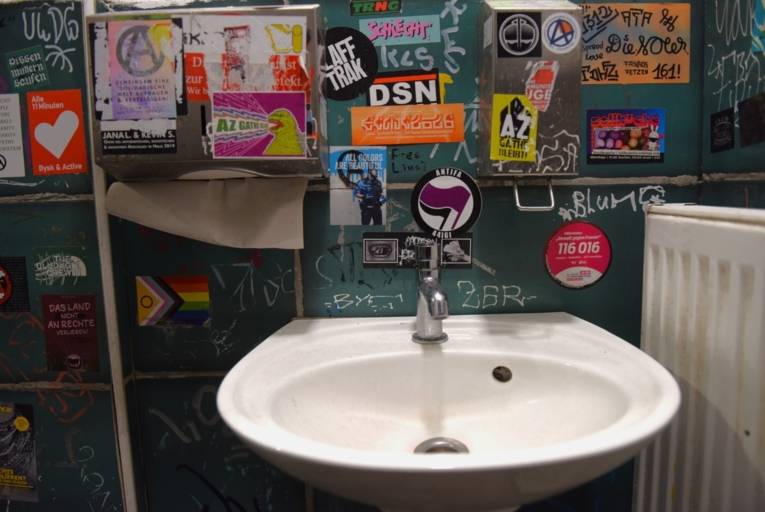 Ein Waschbecken hängt an einer dunklen Wand. Daneben hängt ein Handtuchspender und auf der anderen Seite ist ein Heizkörper. Alles ist mit Stickern beklebt, einige davon mit politischen Statements, pro Demokratie.