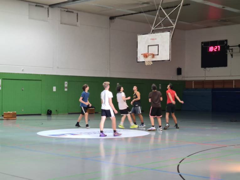 Auf dem Bild sind sechs junge Männer zu sehen, die Basketball spielen in einer Turnhalle.