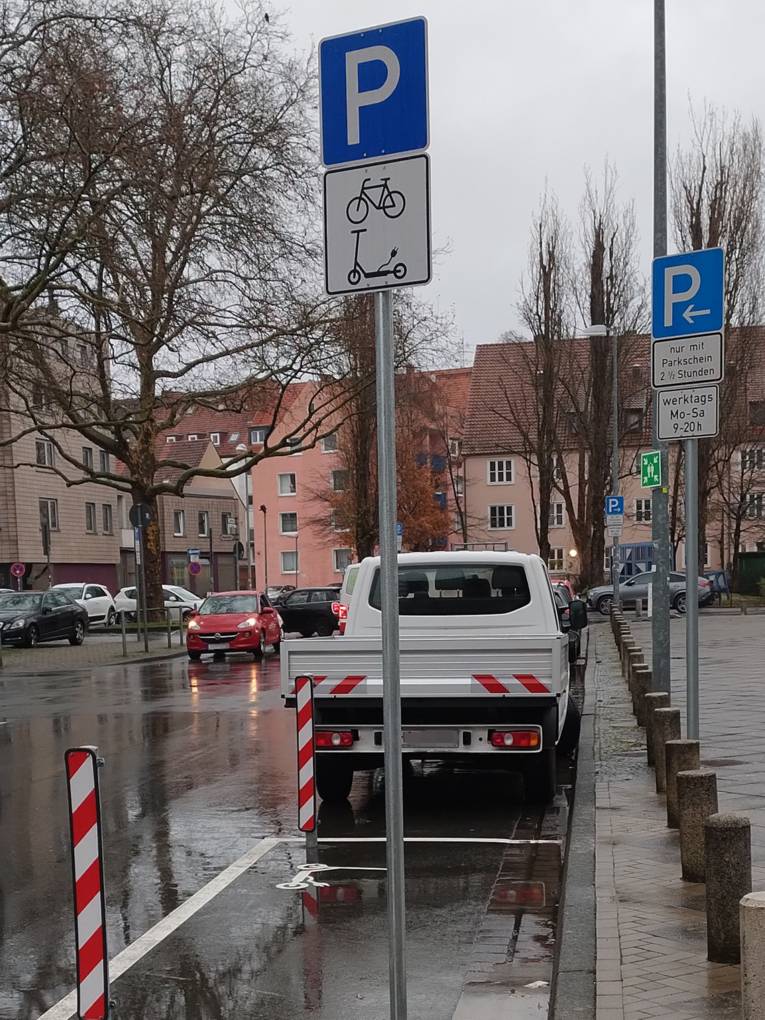 Ein Park-Schild, darunter die Symbole Fahrrad und E-Roller, steht am Stra&szlig;enrand auf einer wei&szlig; markierten Aspaltfl&auml;che. Auf dem Boden ist ein wei&szlig;es Rollersybol mit Stromstecker aufgetragen.