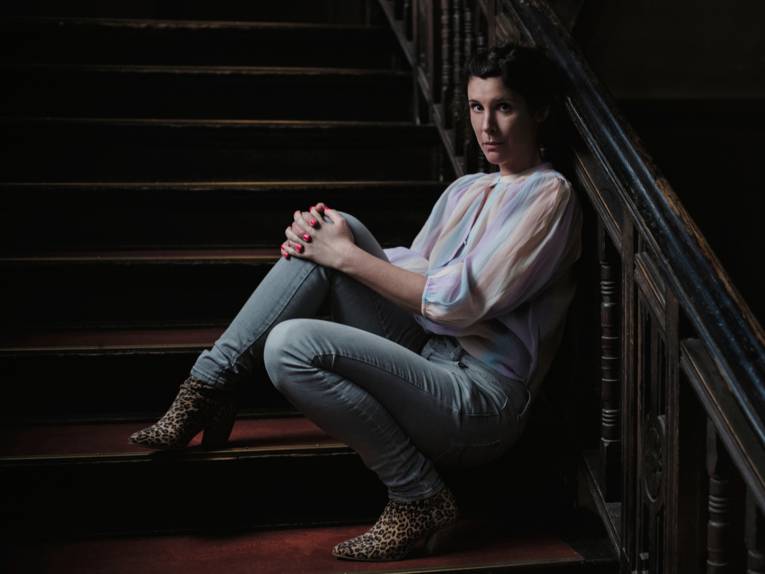 Eine Frau in einer Bluse und Jeans sitzt seitlich auf einer alten, breiten Treppe, dabei lehnt sie gegen das Geländer.