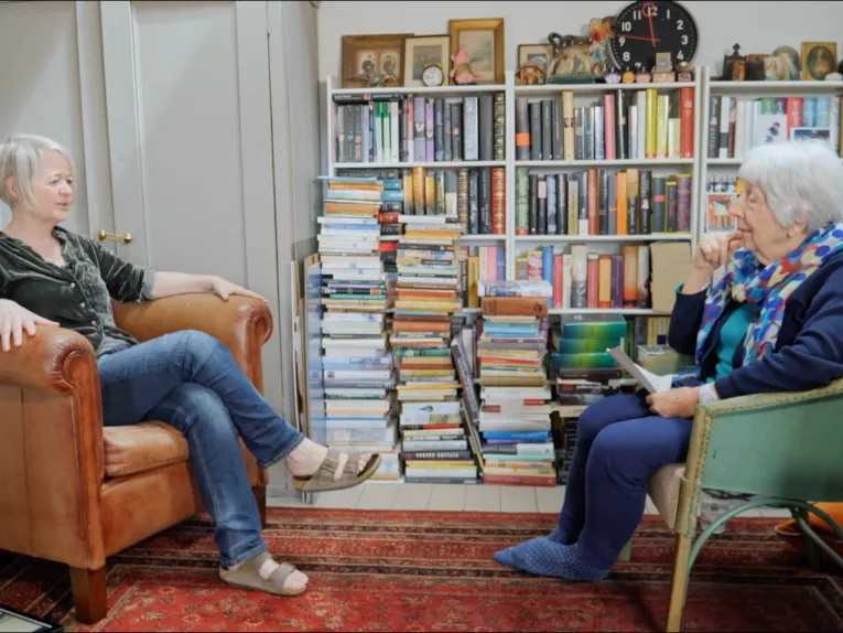 Das Bild zeigt zwei Personen, die sich in einem wohnlichen Raum gegenübersitzen und miteinander sprechen. Im Hintergrund befinden sich gut gefüllte Bücherregale mit zahlreichen Büchern, Bildern und dekorativen Gegenständen. Die Szene wirkt wie ein Interview oder ein persönliches Gespräch in entspannter Atmosphäre. Ein gemusterter Teppich und die gemütlichen Sitzmöbel unterstreichen den privaten, kulturell geprägten Charakter des Raumes.