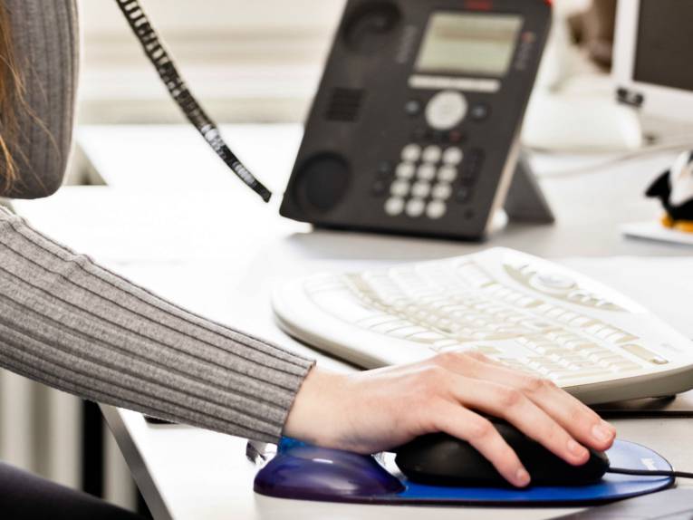 Etwas fragen und Antworten bekommen: Eine Hand bewegt eine Computermaus, vom Telefon ist der Hörer abgenommen und die Person telefoniert mit der anderen Hand.