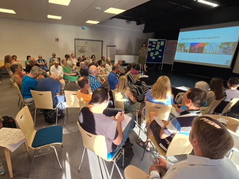 Menschen sitzen auf St&uuml;hlen und blicken nach rechts, wo eine Frau auf einem Podest neben einer Leinwand mit Abbildungen steht.