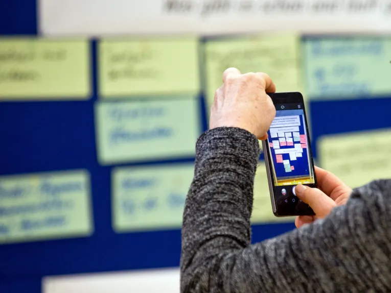 Zwei Hände halten ein Smartphone, das Display zeigt, was die Fotokamera gerade von einer Metaplanwand mit handbeschriebenen Moderationskärtchen aufnimmt.