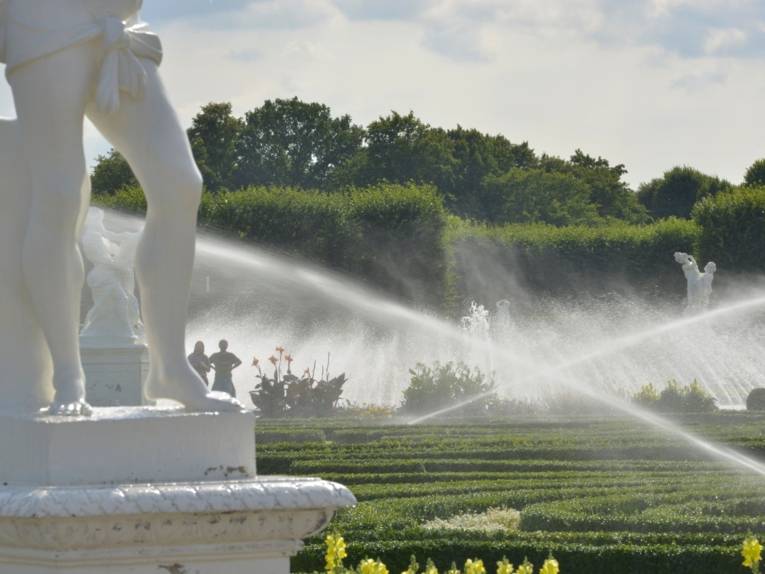 Vorn links die Beine einer weißen Skulptur auf einem Sockel, dahinter zwei überkreuzte Wasserstrahlen, aus denen es auf grüne Linien aus flachen Pflanzen regnet.