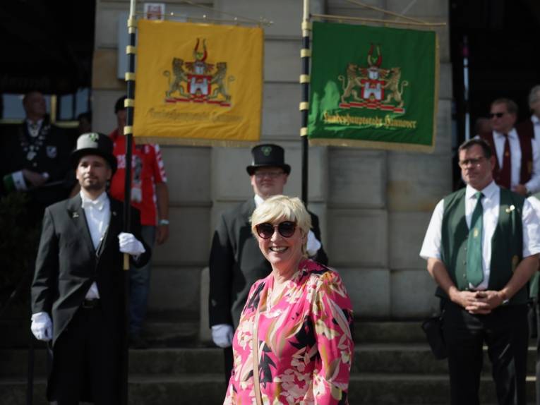 Eine lächelnde Frau in buntem Kleid auf einer Treppe. Im Hintergrund drei Männer in Uniformen.