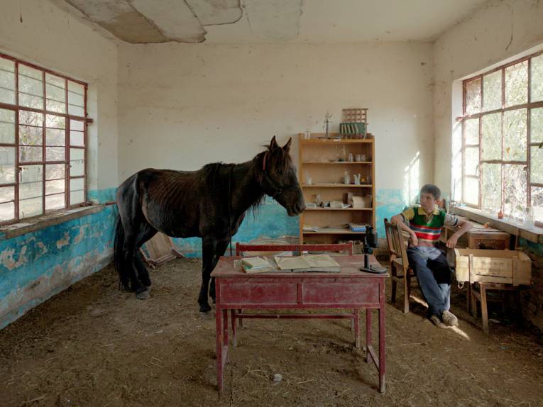 Ein sparsam eingerichter Raum. Auf einem Stuhl sitzt ein Junge. Ein Pferd steht im Raum