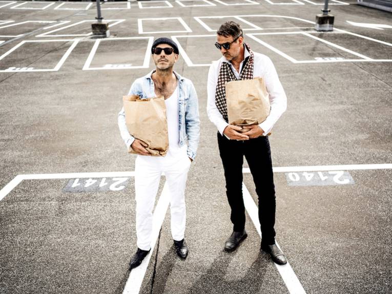 Zwei Männer stehen mit Papiertüten in den Armen auf einem Parkplatz.