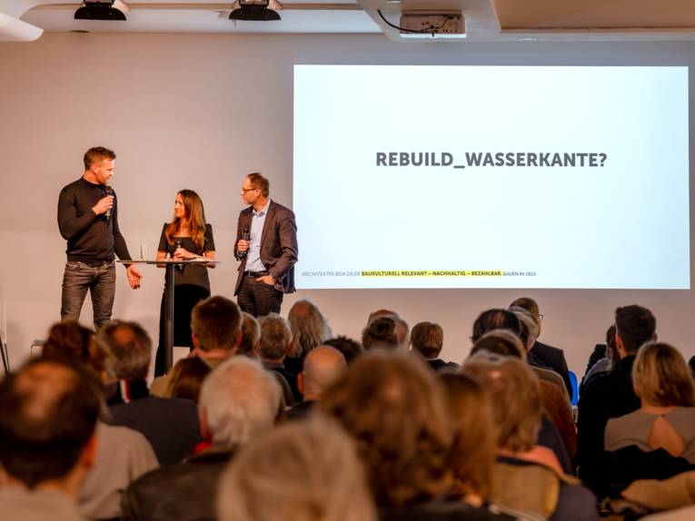 Ein Publikum von hinten sieht auf eine Plattform, auf der drei Personen an einem Stehtisch mit Mikrofonen stehen, rechts daneben eine auf die Wand projezierte Seite mit "Rebuilt_Wasserkante".