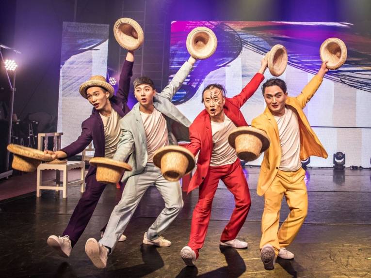 Zu sehen sind vier chinesische Männer in verschiedenfarbigen Anzügen auf der Bühne agierend.