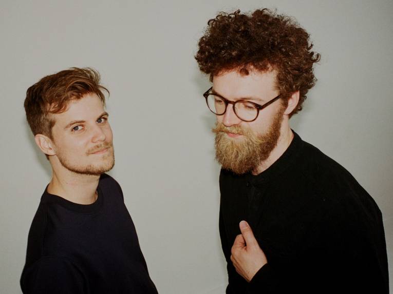 Ein Mann mit kurzem Haar und Fünftagebart und einer mit kurzen Locken, Brille und Vollbart