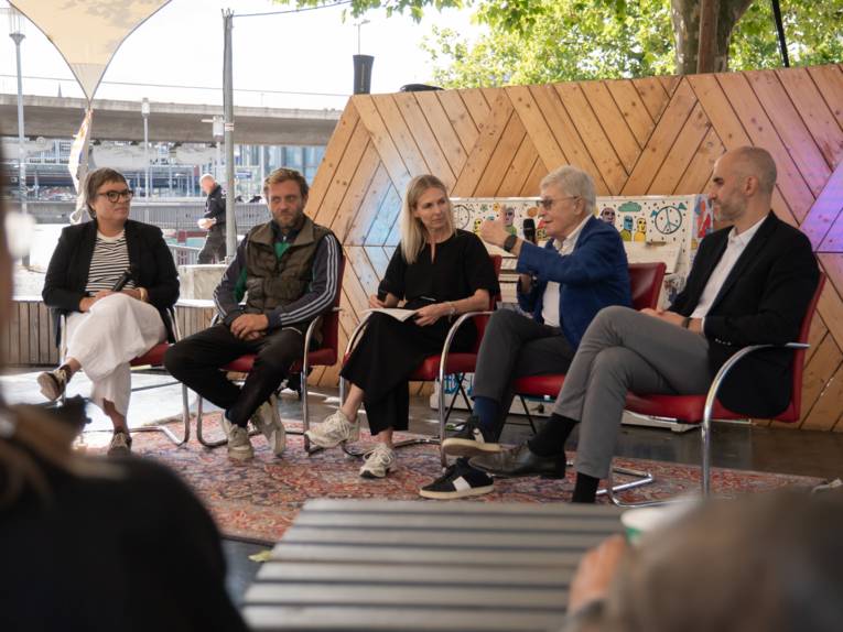 Vier Menschen sitzen auf Stühlen auf einer kleinen Bühne, ein Mann spricht in ein Mikrofon.