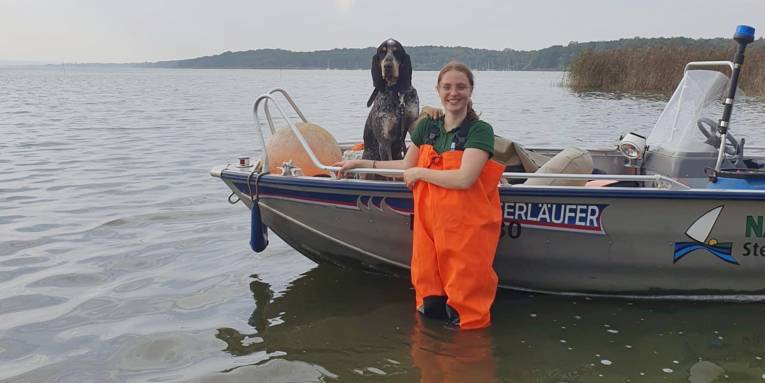 Eine Frau tr&auml;gt eine orangefarbene Wathose und steht vor einem Boot im Wasser des Steinhuder Meeres. Im Boot sitzt ein Hund und schaut mit der Frau in die Kamera.