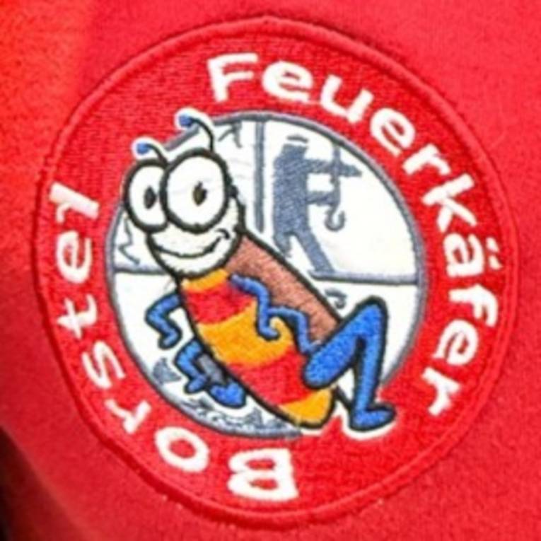 Aufn&auml;her auf einem roten Kleidungsst&uuml;ck: In der Mitte ist ein K&auml;fer, hinter ihm ist das Feuerwehr-Emblem, das die vier Hauptaufgaben der Feuerwehr darstellt: Retten / L&ouml;schen / Bergen / Sch&uuml;tzen. Drumherum steht kreisrund: "Feuerk&auml;fer Borstel".