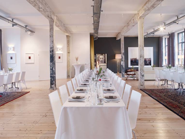 Stilvolle Eventlocation "Helmkehof" mit langen, festlich gedeckten Tischen, rustikalen Säulen und moderner Kunst an den Wänden.