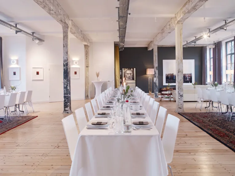 Stilvolle Eventlocation "Helmkehof" mit langen, festlich gedeckten Tischen, rustikalen Säulen und moderner Kunst an den Wänden.
