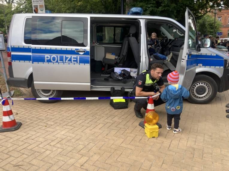 Einsatzfahrzeug und Polizist, der mit einem Kleinkind spricht. Er ist in der Hocke.