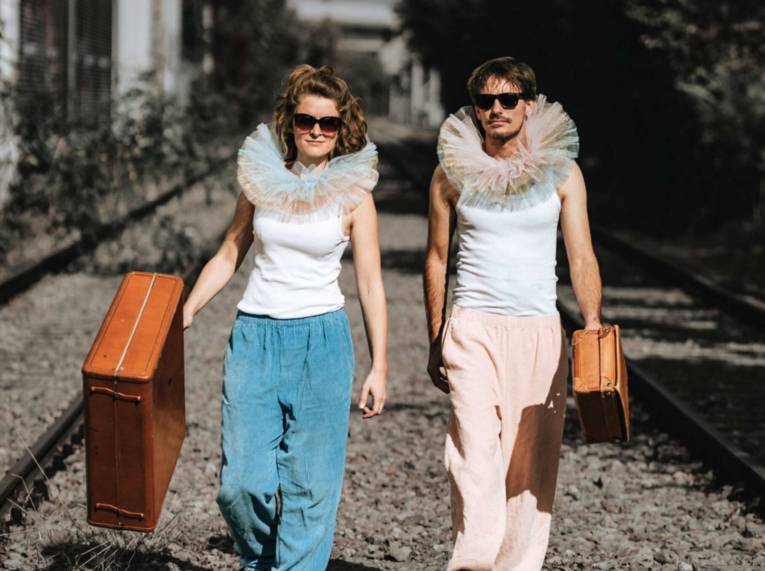 Ein Mann und eine Frau mit Sonnenbrillen und großen Halskrausen aus Tüll laufen mit Koffern zwischen Bahngleisen.
