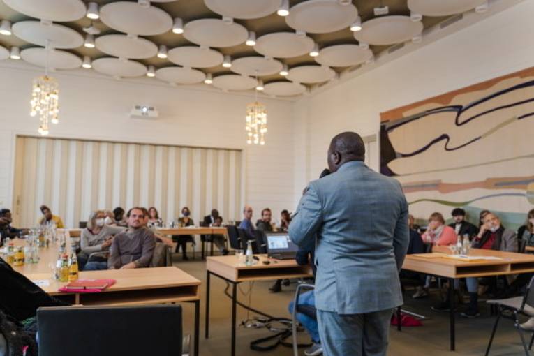 Mehrere sitzende Personen in einem Raum, die einem Vortragenden zuhören.