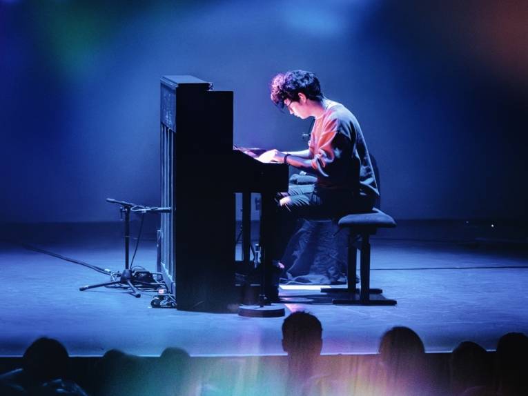 Zu sehen ist der Pianist Tony Ann. Er spielt an einem schlichten Klavier, das auf der Bühne steht.