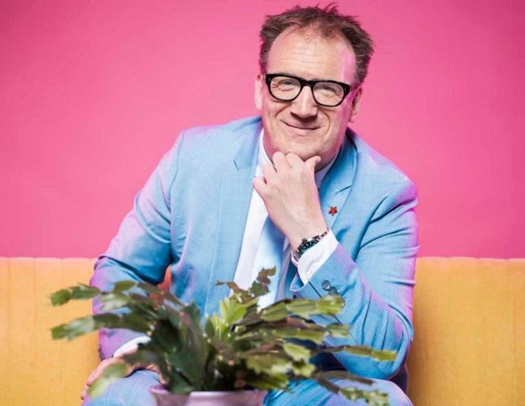 Lächelnder Mann mit kurzem Haar, und Brille in Hemd und Jackett, vor ihm steht eine Topfpflanze.