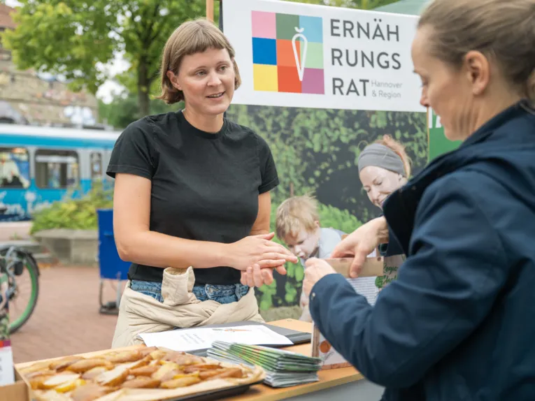 Eine Frau hinter einem Tisch redet mit einer anderen Frau ihr gegen&uuml;ber. Hinter der ersten steht ein Schild mit der Aufschrift "Ern&auml;hrungsrat Hannover".