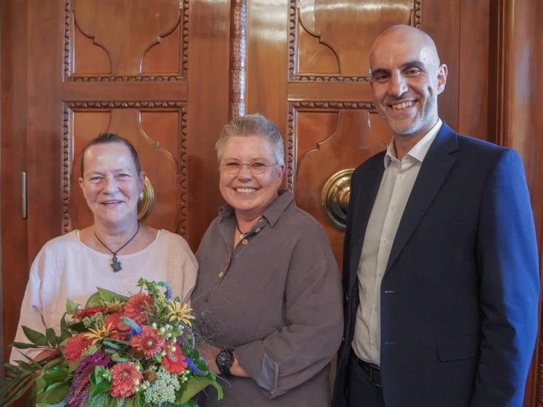 Auf dem Bild sind zwei Frauen und ein Mann zu sehen, die nebeneinander stehen. Die Frau ganz links hält einen Blumen